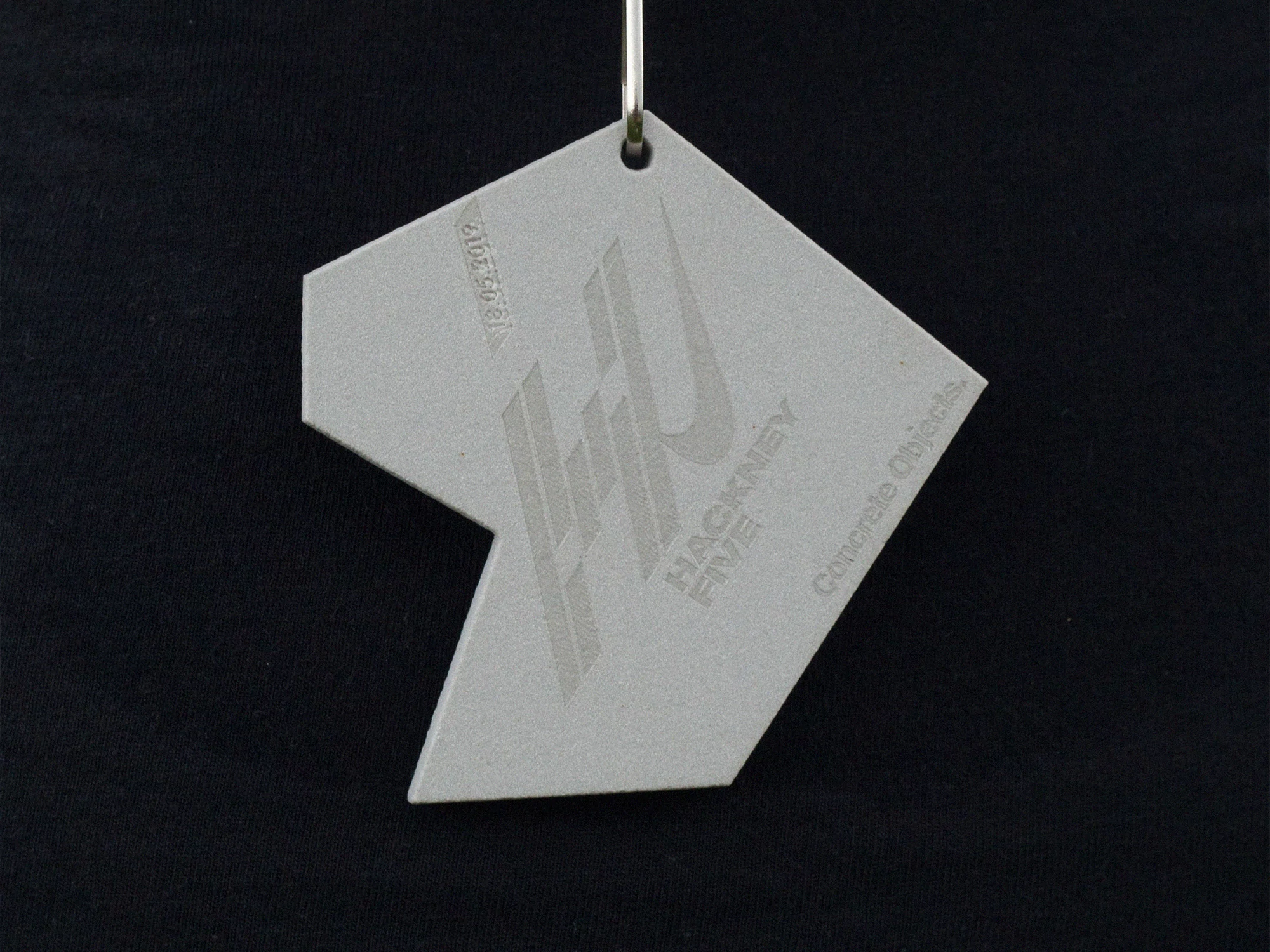 Close-up of a geometric gray tag with a design and text, hanging from a metal ring against a black background. Minimalist design.