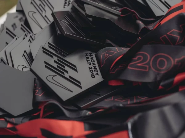 Close-up of stacked Hackney Half 2019 participation medals with Nike swoosh and patterned fabric ribbons, showcasing race memorabilia.