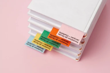 Stack of white books with colourful bookmarks featuring braille and text on a pink surface