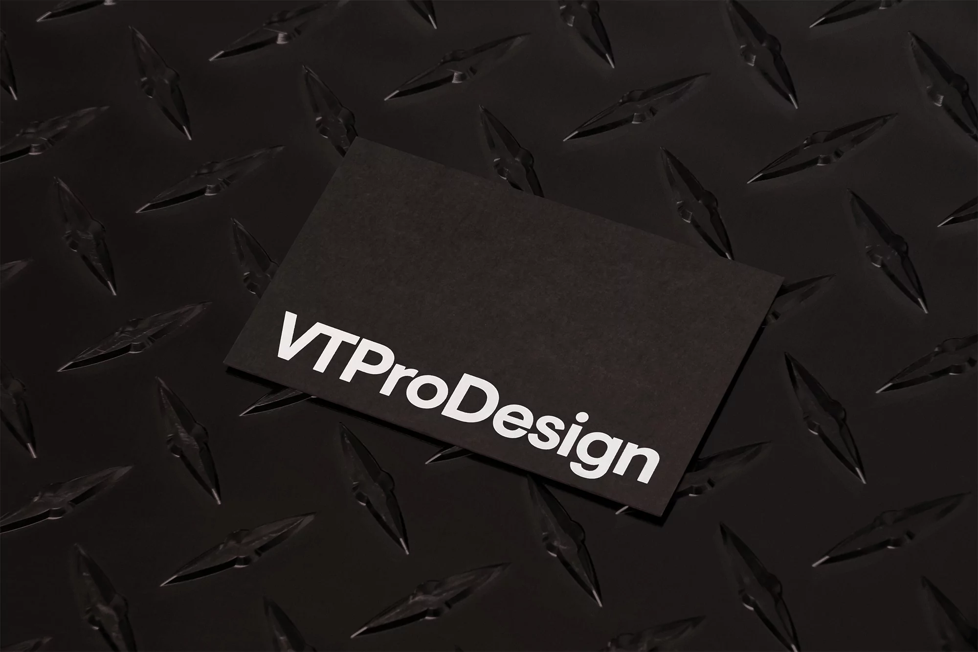 A black business card with white text, VTProDesign, sits atop a textured, dark, industrial surface with a repeating diamond pattern.