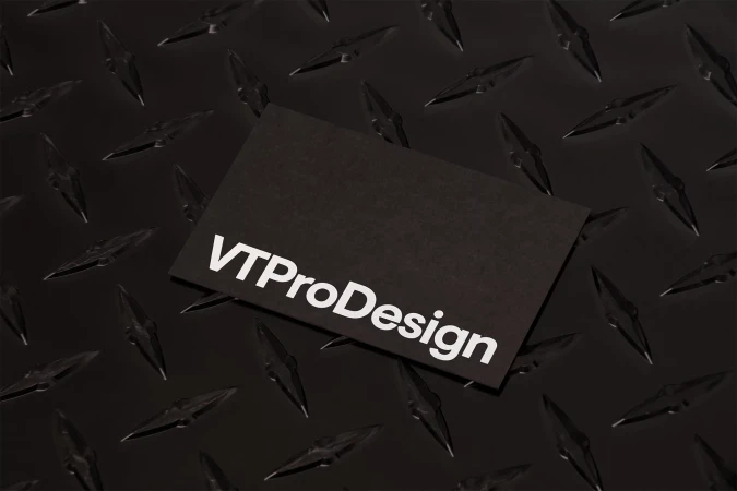 A black business card with white text, VTProDesign, sits atop a textured, dark, industrial surface with a repeating diamond pattern.