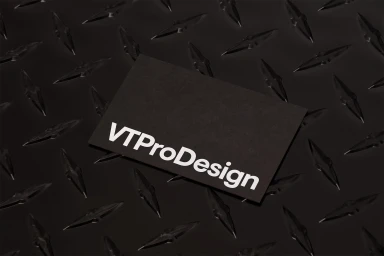 Black business card with white VTProDesign typography resting on a dark metal treadplate surface.