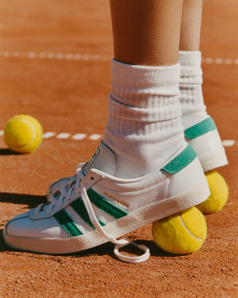 Close-up of white Adidas sneakers with green stripes on tennis court, balanced on two yellow tennis balls. Focus on footwear and sport.