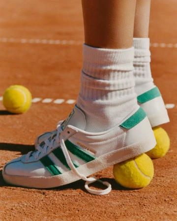 Close-up of white Adidas sneakers with green stripes on tennis court, balanced on two yellow tennis balls. Focus on footwear and sport.
