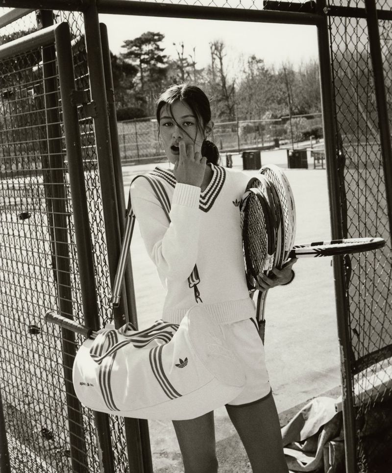 A woman in tennis whites stands on a tennis court holding tennis rackets and a sports bag, posing in a monochrome photograph.
