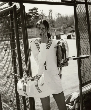 A woman in tennis whites stands on a tennis court holding tennis rackets and a sports bag, posing in a monochrome photograph.