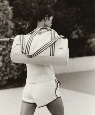 Back view of an adult female in tennis apparel, carrying an Adidas sports bag and holding a tennis racket, with a blurred outdoor backdrop.