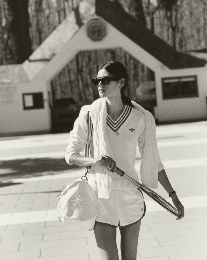 Woman in a white tennis outfit carrying a racket and duffel bag in front of a building and trees, shot in black and white.