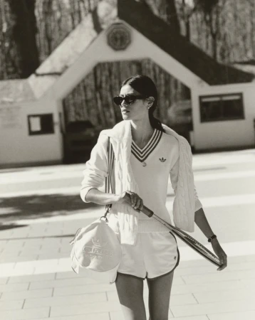 Woman in a white tennis outfit carrying a racket and duffel bag in front of a building and trees, shot in black and white.