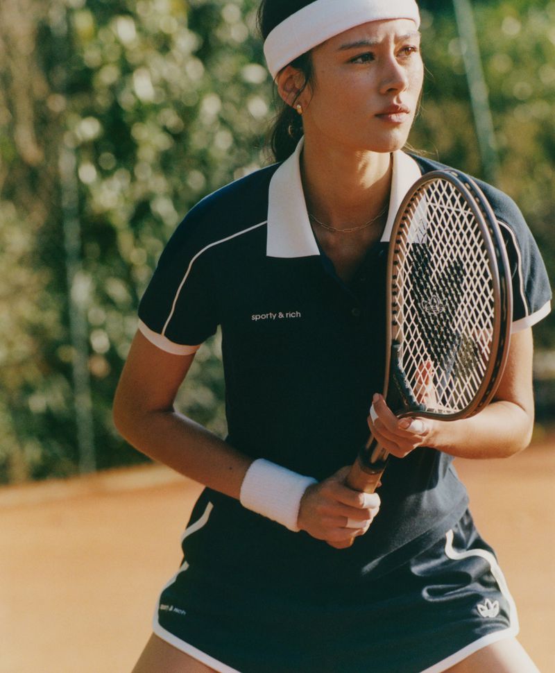Woman in sportswear holds a tennis racket on a clay court. She wears a headband, wristband, and a 'Sporty & Rich' branded athletic outfit.