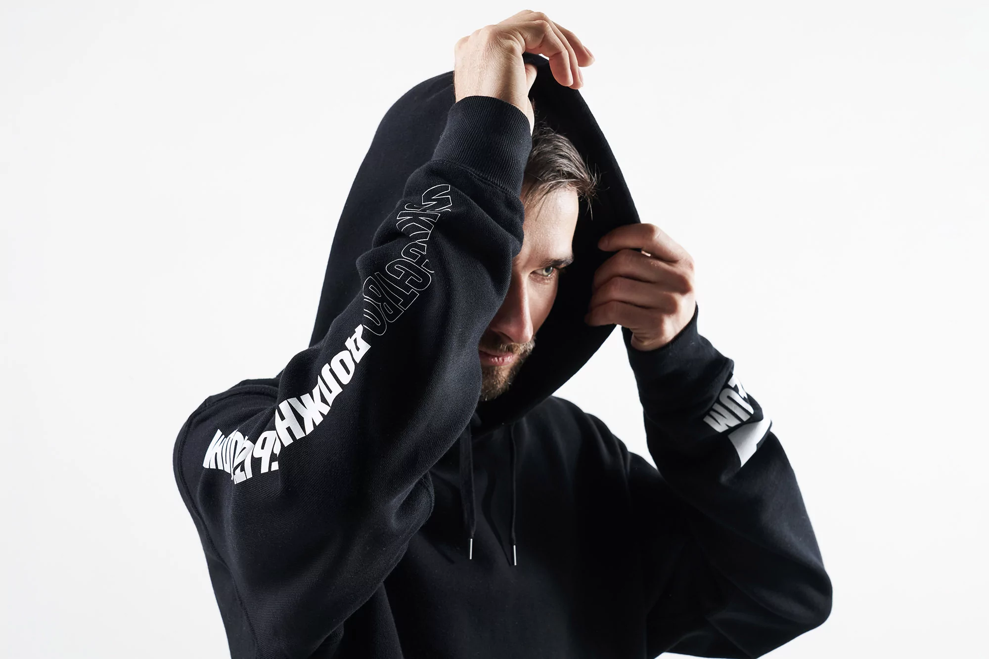 A bearded man wearing a black hooded sweatshirt adjusts the hood against a white backdrop in a studio setting.