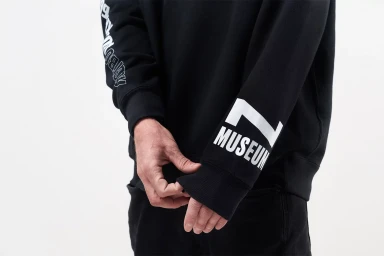 Close-up of a person wearing a black sweatshirt with white typography reading MUSEUM on the sleeve