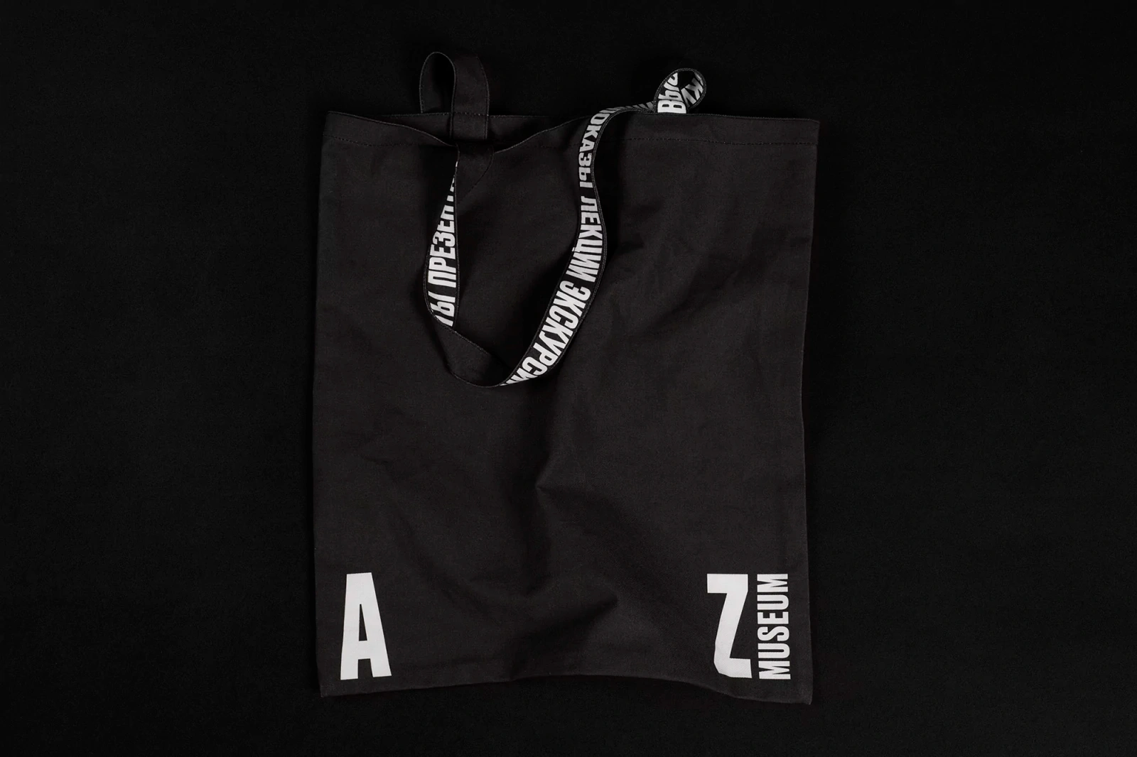 Black canvas tote bag with white typography straps and museum logo resting on a dark background