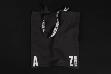 Black canvas tote bag with white typography straps and museum logo resting on a dark background