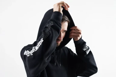 Man wearing a black hoodie with white typography on the sleeve, holding the hood up against a white background.