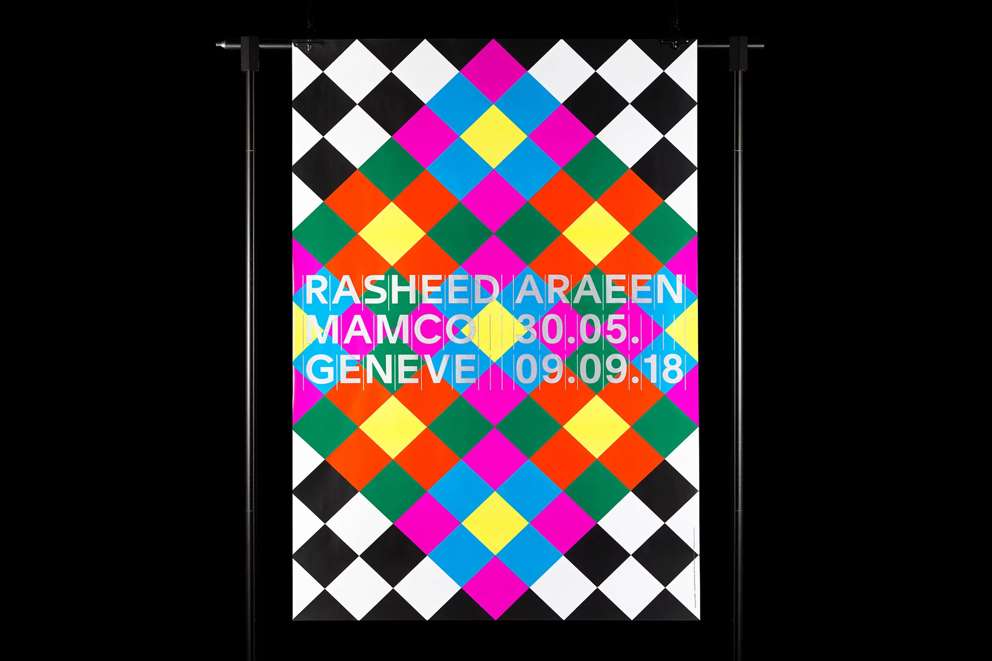 Abstract poster with geometric diamond pattern in black, white, yellow, pink, green, blue, and orange on a black backdrop, hanging on a stand.