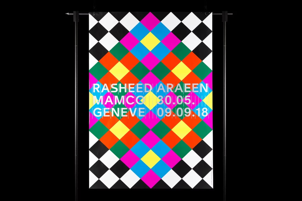 Abstract poster with geometric diamond pattern in black, white, yellow, pink, green, blue, and orange on a black backdrop, hanging on a stand.