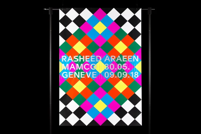 Abstract poster with geometric diamond pattern in black, white, yellow, pink, green, blue, and orange on a black backdrop, hanging on a stand.