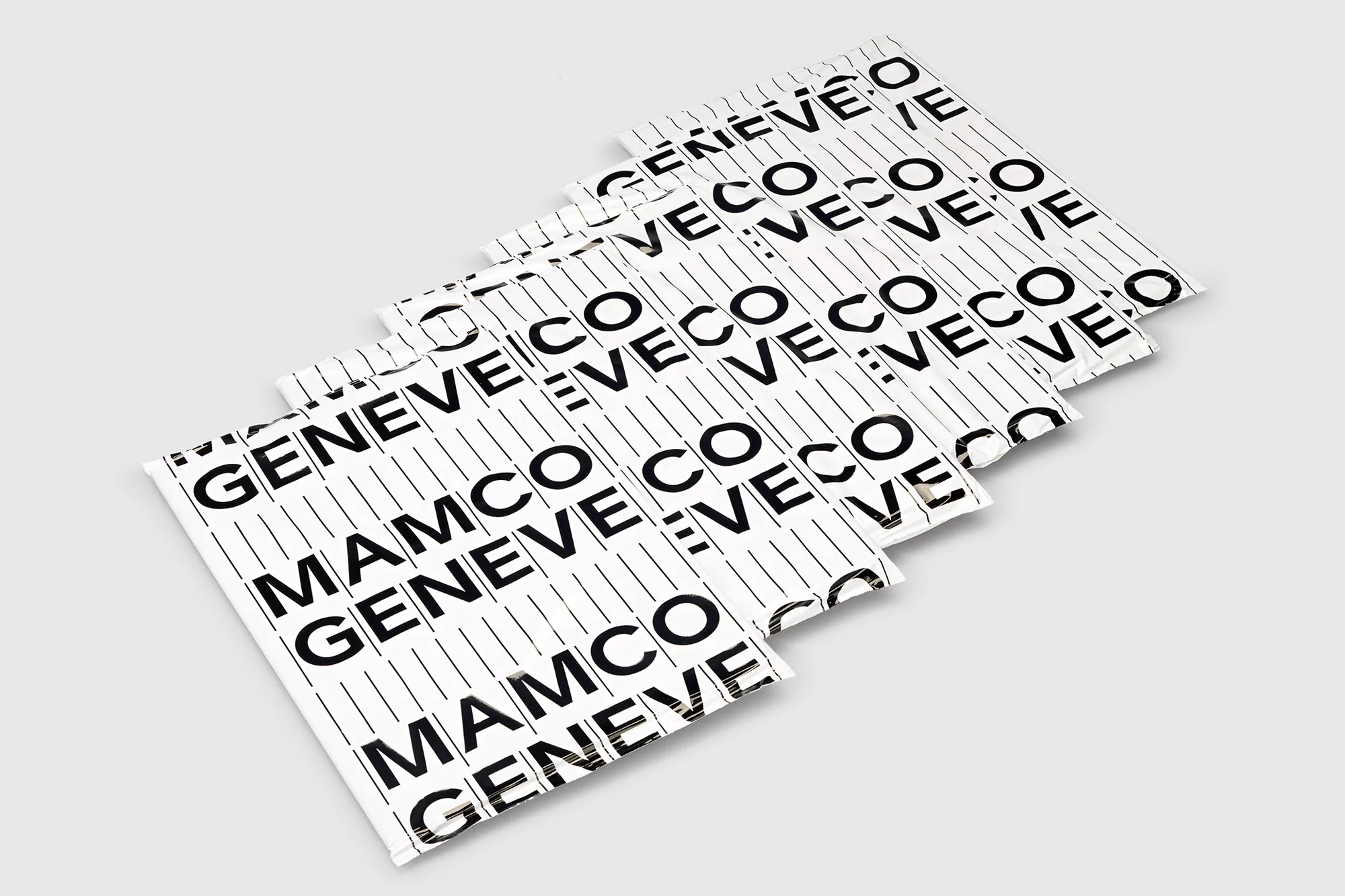 Multiple white envelopes with the repeating text 'MAMCO GENEVE' in bold black font, stacked diagonally on a solid light gray surface.