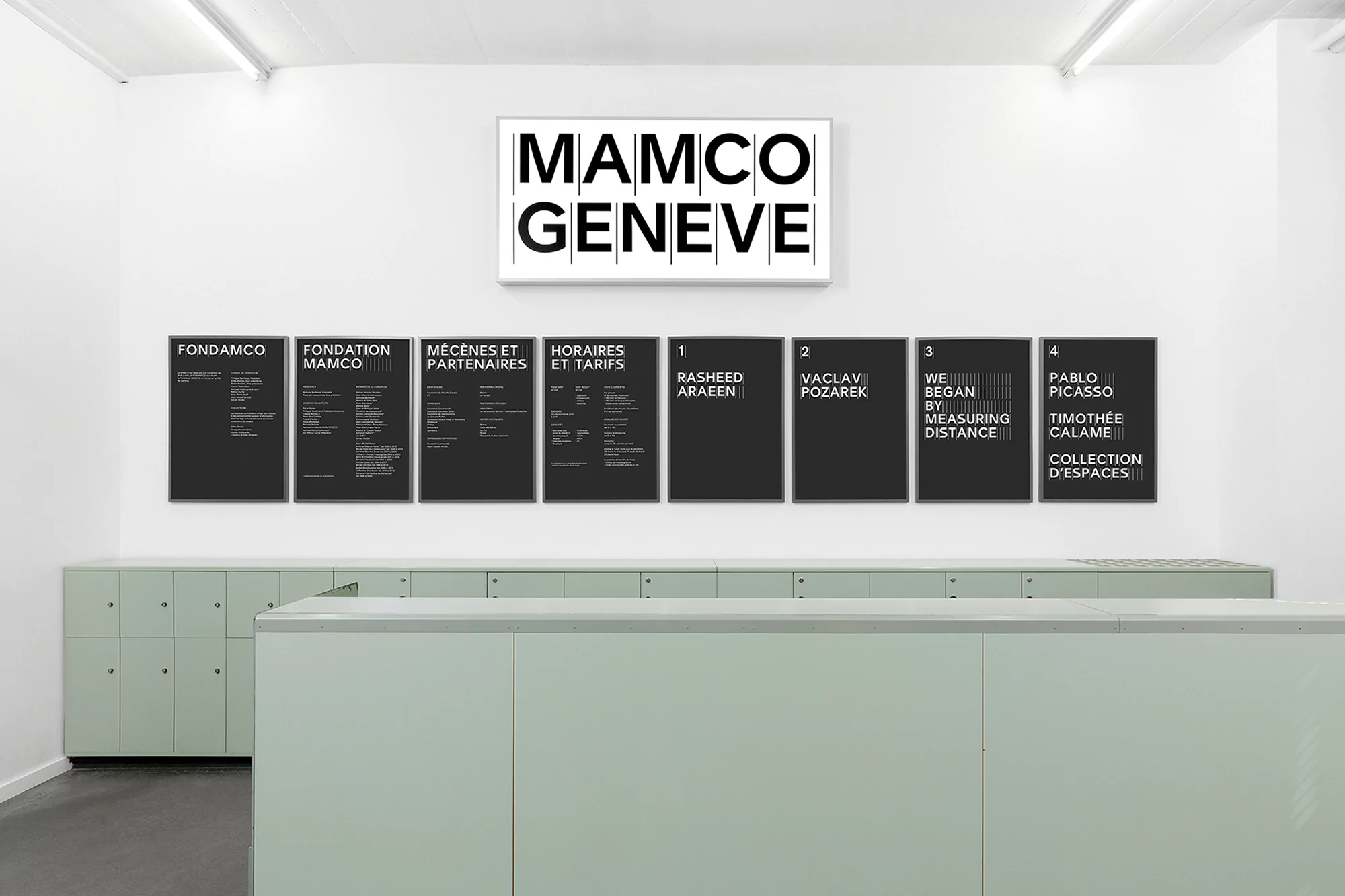 Exhibition view featuring MAMCO Geneve banner and posters with exhibit titles in a minimalist, brightly lit, white-walled gallery space.