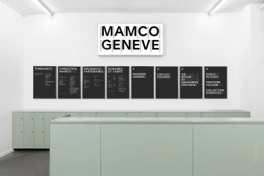 Mamco Geneve museum interior showcasing black informational posters and a prominent brand lightbox on a white wall.