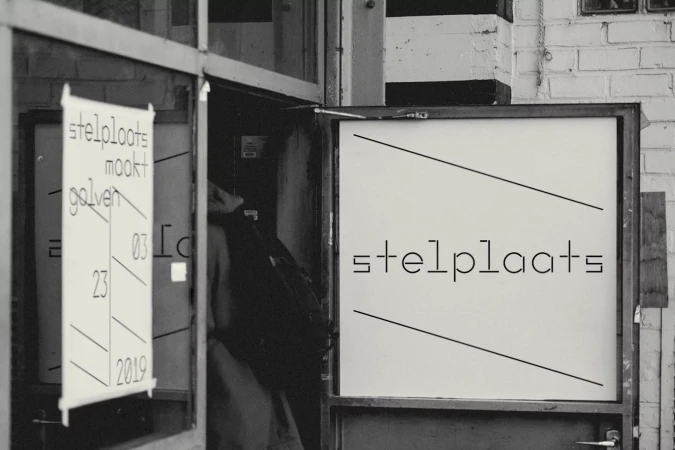 Black and white image of a person entering an art space, with posters advertising 'stelplaats maakt golven' and event details.