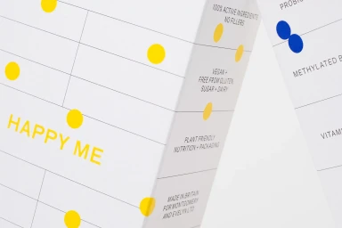 Close-up of white packaging with yellow and blue circular dots, grid lines, and text including HAPPY ME for Montgomery+Evelyn.