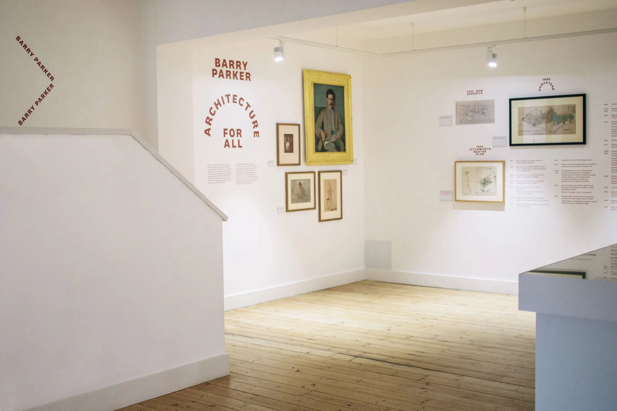 Interior view of an art gallery exhibition with artwork displayed on white walls and a light wood floor, featuring architectural design.