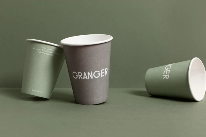 Three paper cups with white lids rest on a green surface against a green background; one cup displays the name 'GRANGER' in white letters.