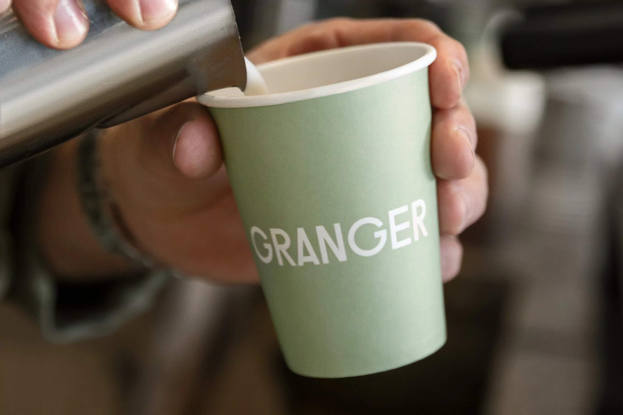 Close-up shot of milk being poured from a metal pitcher into a light green disposable coffee cup, held by a hand, indoors.