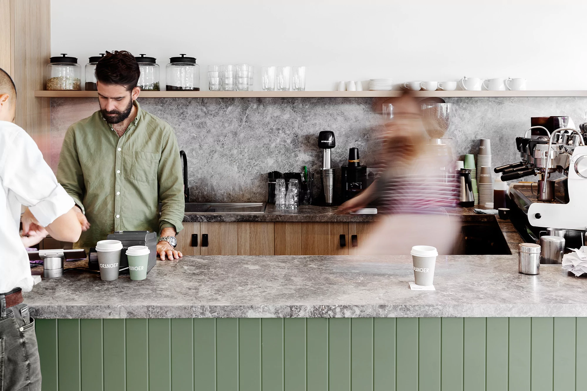 Bearded barista serves customer at cafe counter. Coffee cups, espresso machine, and neutral color palette create a modern coffee shop scene.