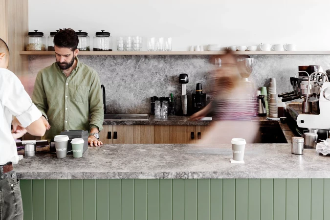 Bearded barista serves customer at cafe counter. Coffee cups, espresso machine, and neutral color palette create a modern coffee shop scene.