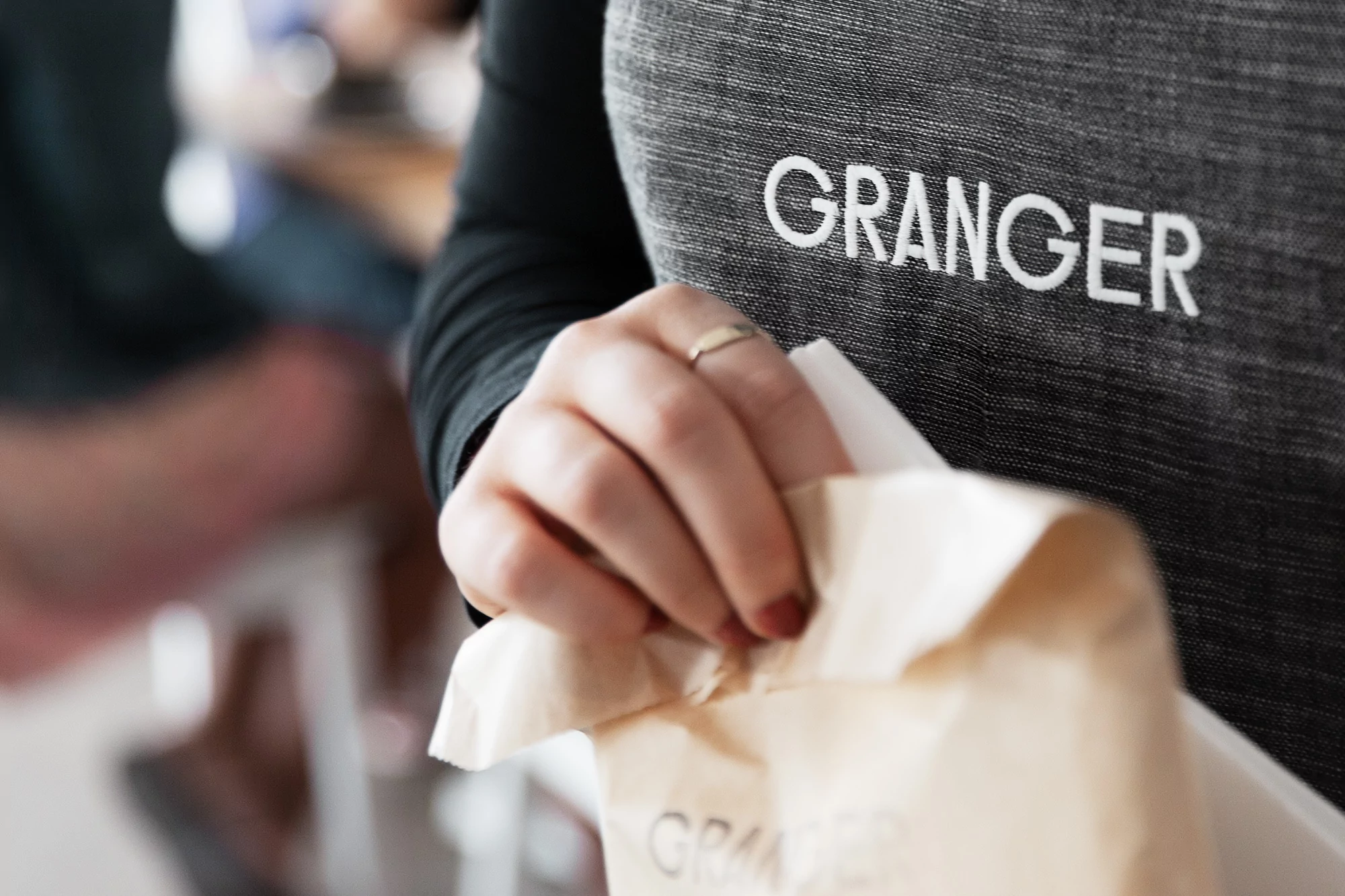 Close-up shot of a person in a gray apron labeled 'GRANGER' holding a paper bag, focus on hands and brand, indoor setting.
