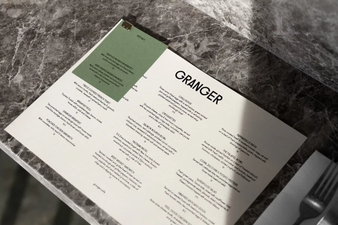 Granger restaurant menu displayed on a marble table, featuring food items like chilli eggs, croquettes, and the falafel bowl.