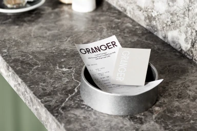 Branded Granger cafe receipt and card resting inside a round metal container on a dark marble countertop.