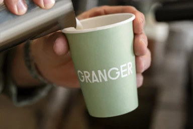 Close-up of a person pouring milk into a light green Granger paper coffee cup with white branding.