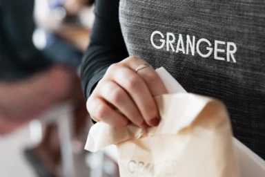 Close-up of a person wearing a textured grey apron with the text GRANGER holding a paper bag.