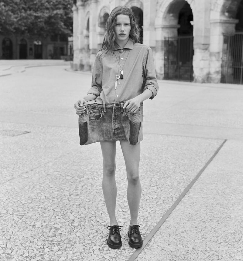 Black and white portrait of a woman in a button-down shirt holding a denim skirt in front of a stone building on a sunny day.