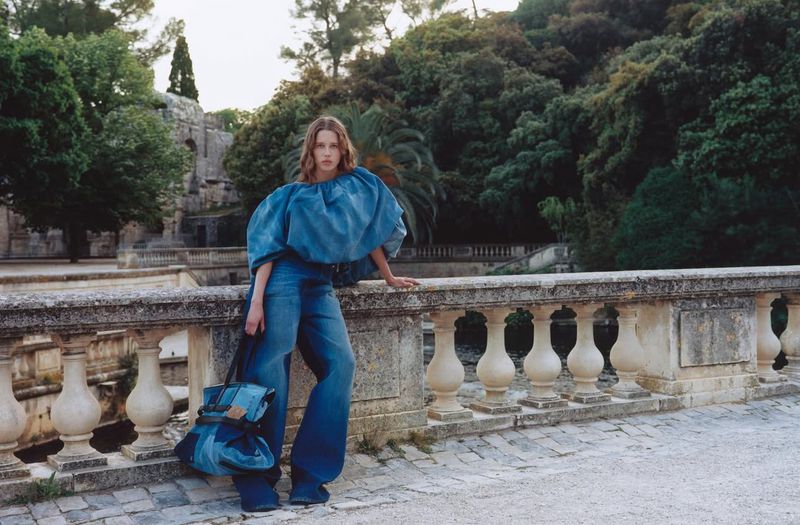 A young adult woman in a denim outfit poses on a stone ledge with a bag, with greenery and architecture in the outdoor background.