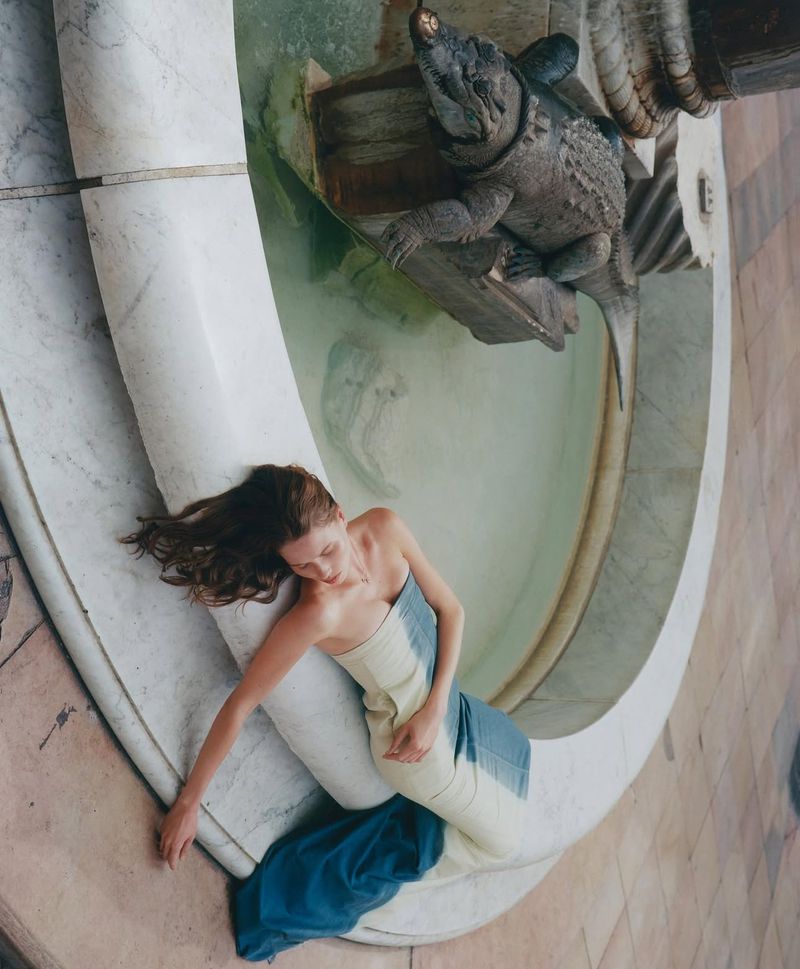 Overhead shot of a woman in a draped white and blue dress lying on a marble fountain with a turtle sculpture and flowing water.