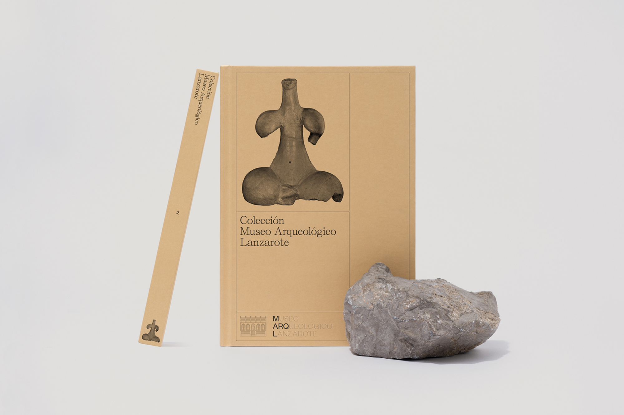 Book titled Colección Museo Arqueológico Lanzarote with an image of a figurine, and a gray rock on a white surface in a studio.