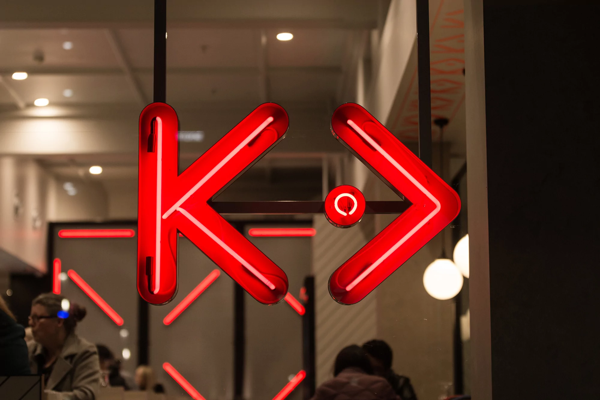 Red neon sign with a stylized 'K' and arrow design hanging in a window, people visible in the background of an indoor setting.
