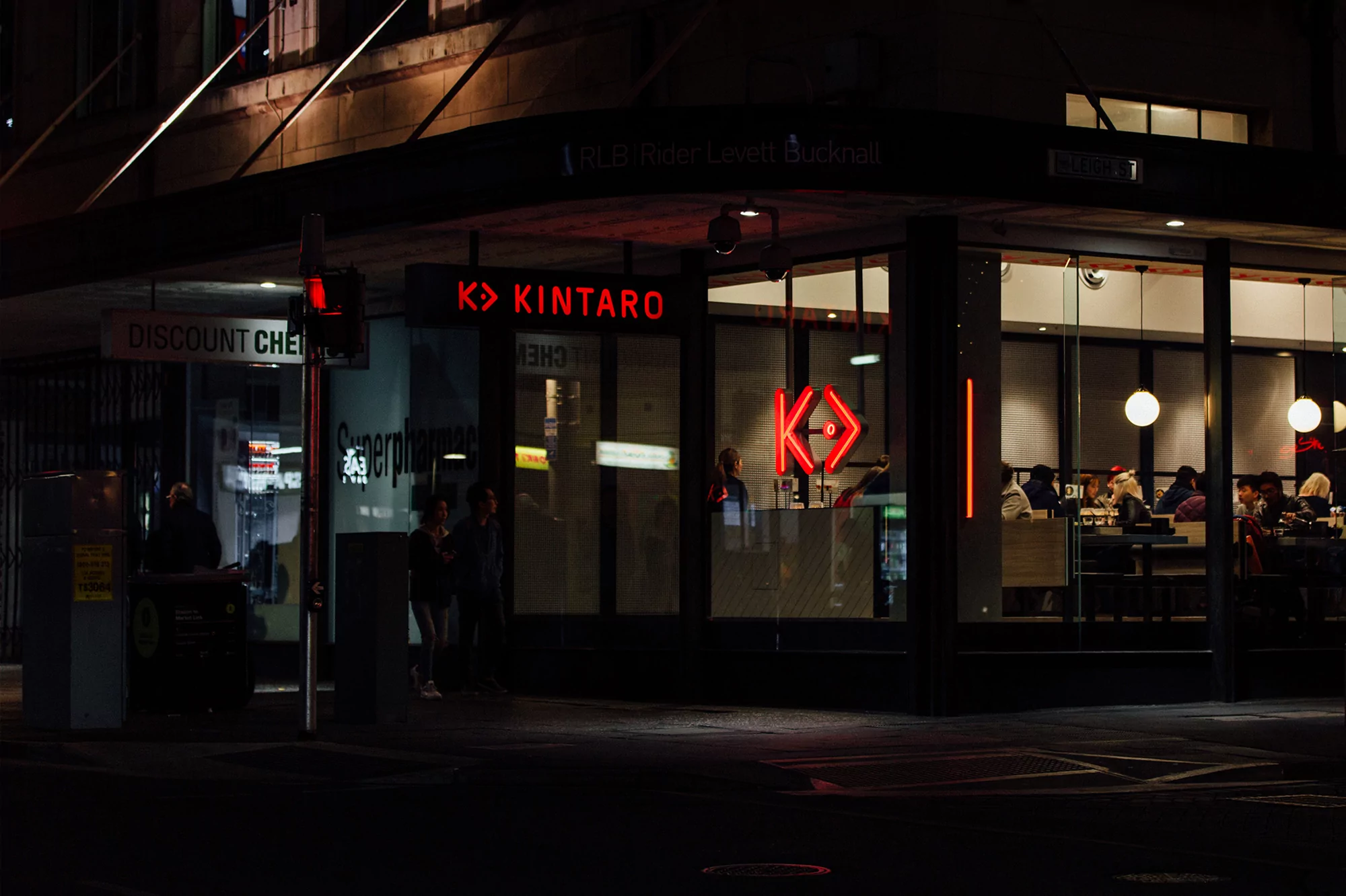 Night view of Kintaro restaurant in urban setting, with people dining visible through the large storefront windows, warmly lit.