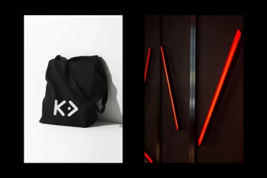 Black canvas tote bag featuring a white logo sitting on a white surface next to a panel with diagonal red neon lights.