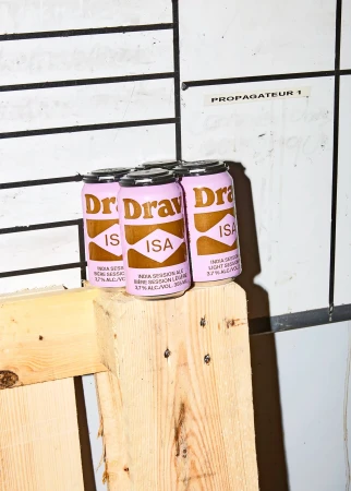 Four cans of Dr. Drava ISA beer are stacked on top of a wooden pallet against a white tiled wall in an indoor setting.
