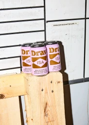 Four pink and gold Drav beer cans resting on a wooden pallet against a white tiled wall with grid lines.