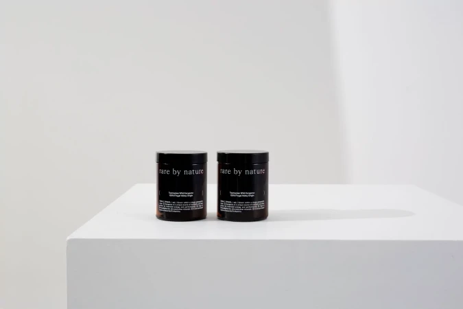 Two dark brown cosmetic jars with 'rare by nature' labels sit on a white surface against a bright white background, product photography.
