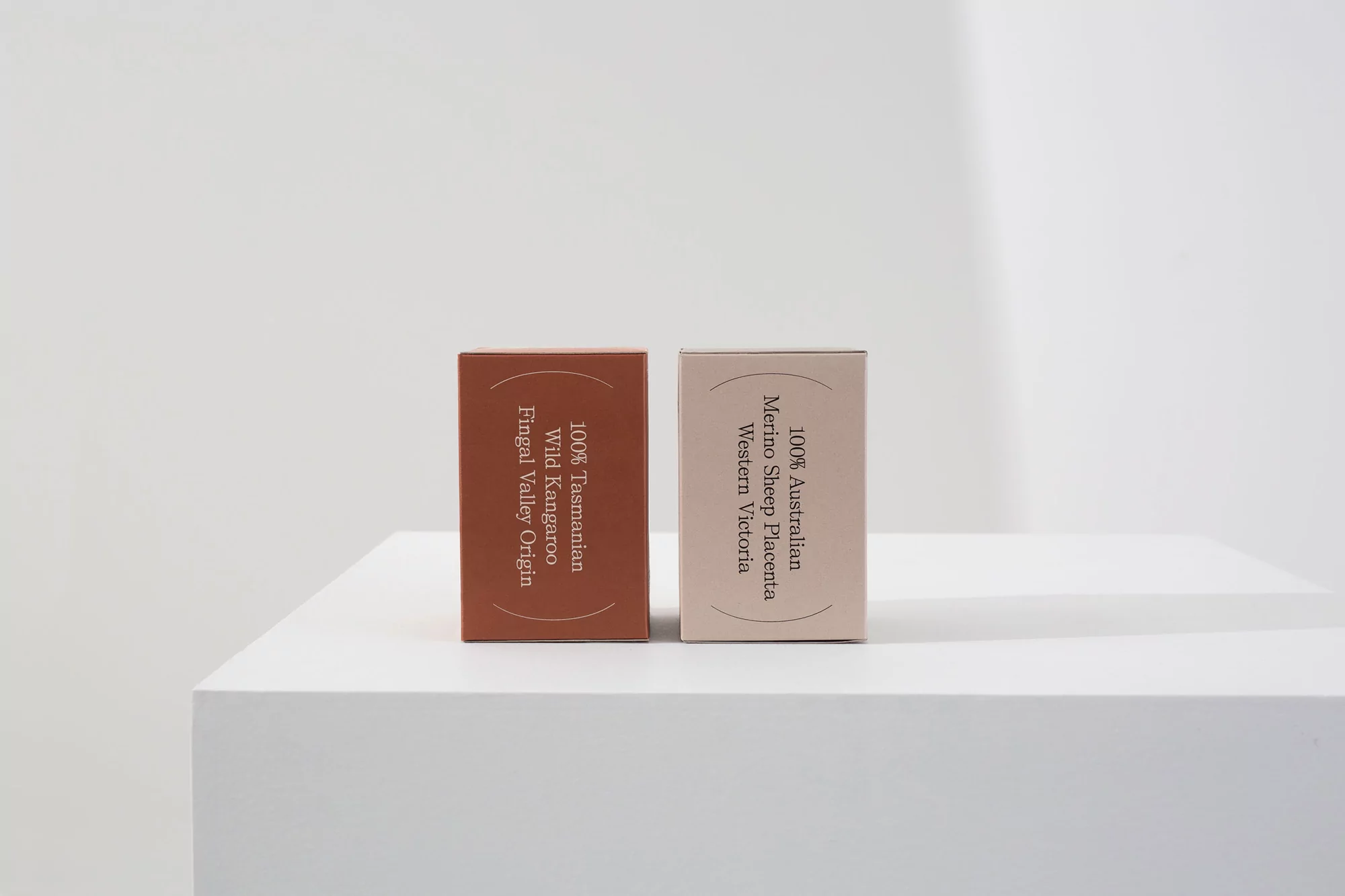 Two boxes of skincare product in neutral tones, one brownish and one light pink, displayed on a white surface in a studio setting.