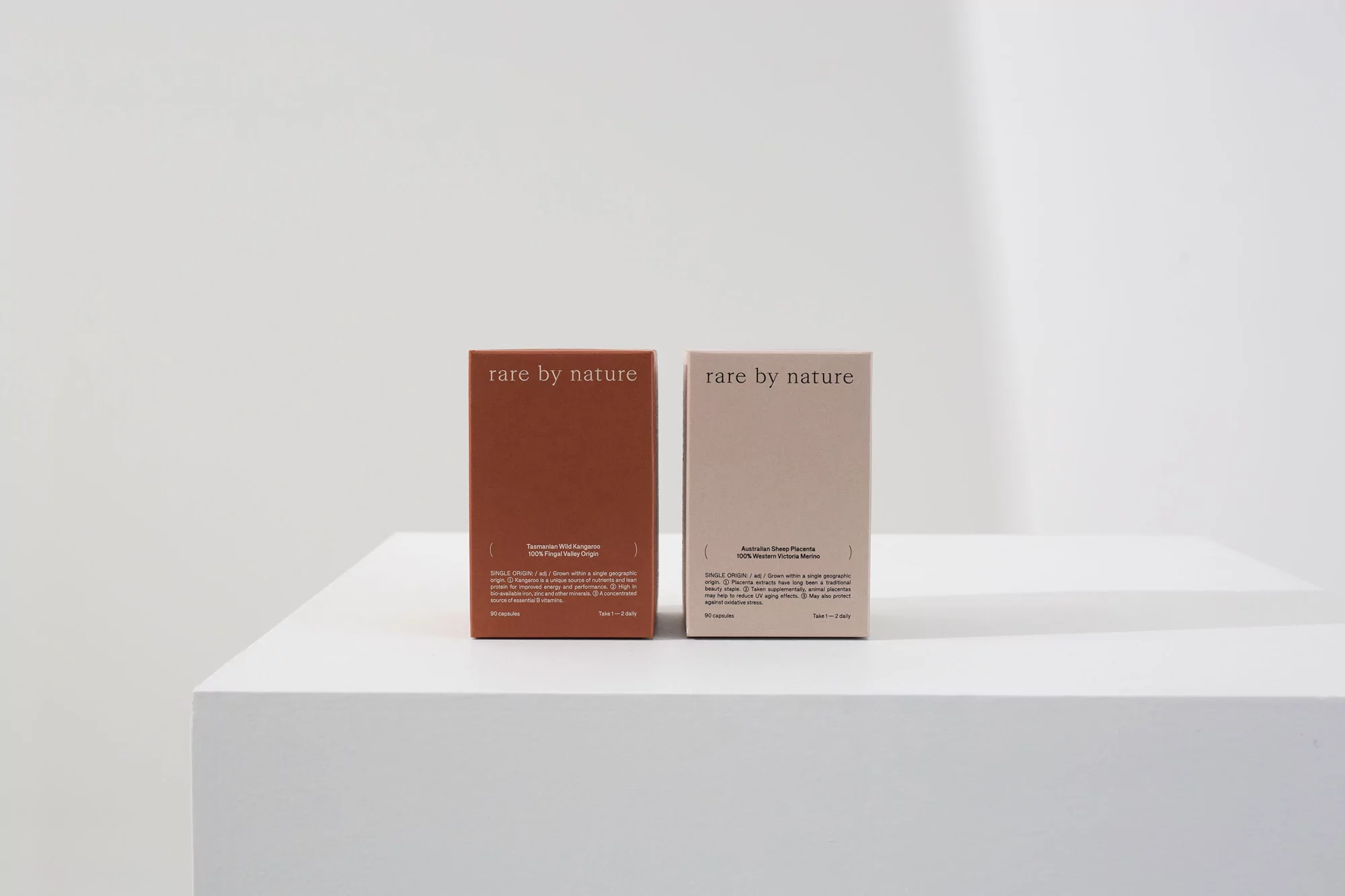 Two 'Rare by Nature' skincare product boxes, one brown and one light beige, stand on a white platform against a bright white backdrop.