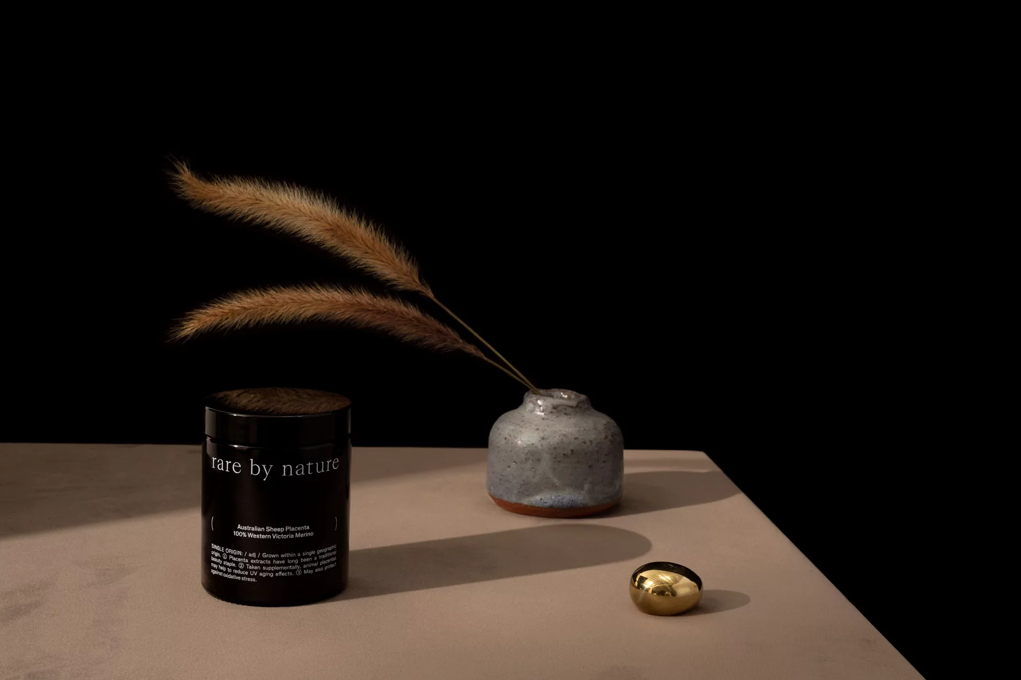 Still life featuring a black cosmetic jar, small grey vase with ornamental grass, and a golden stone on a beige surface against a black backdrop.